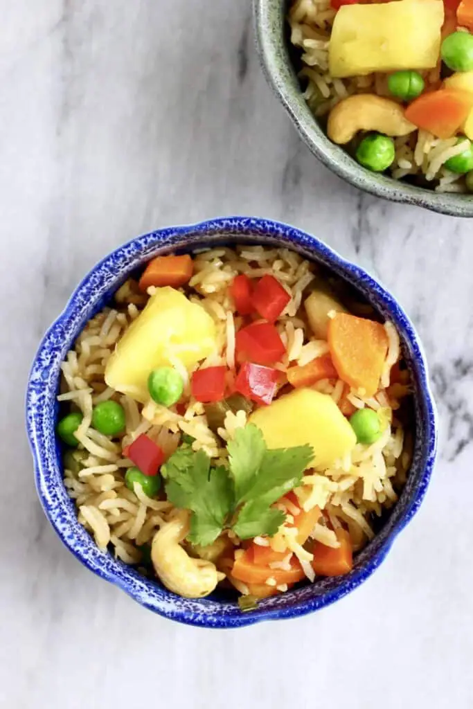 Photo of a small blue bowl with rice, green peas, red pepper, carrots and pineapple chunks against a marble background