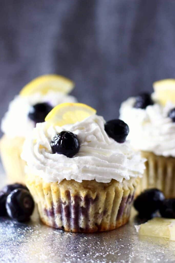 Photo of three blueberry cupcakes topped with white creamy frosting and a fresh blueberry and lemon wedge against a grey background