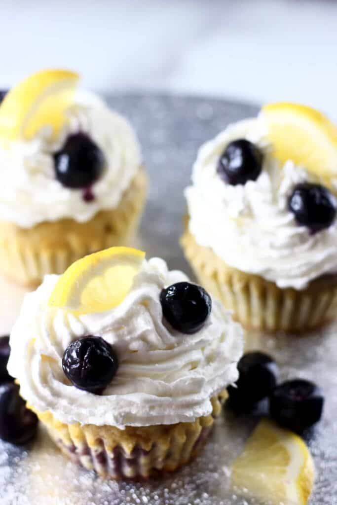 Photo of three cupcakes topped with white creamy frosting, fresh blueberries and lemon wedges on a silver background