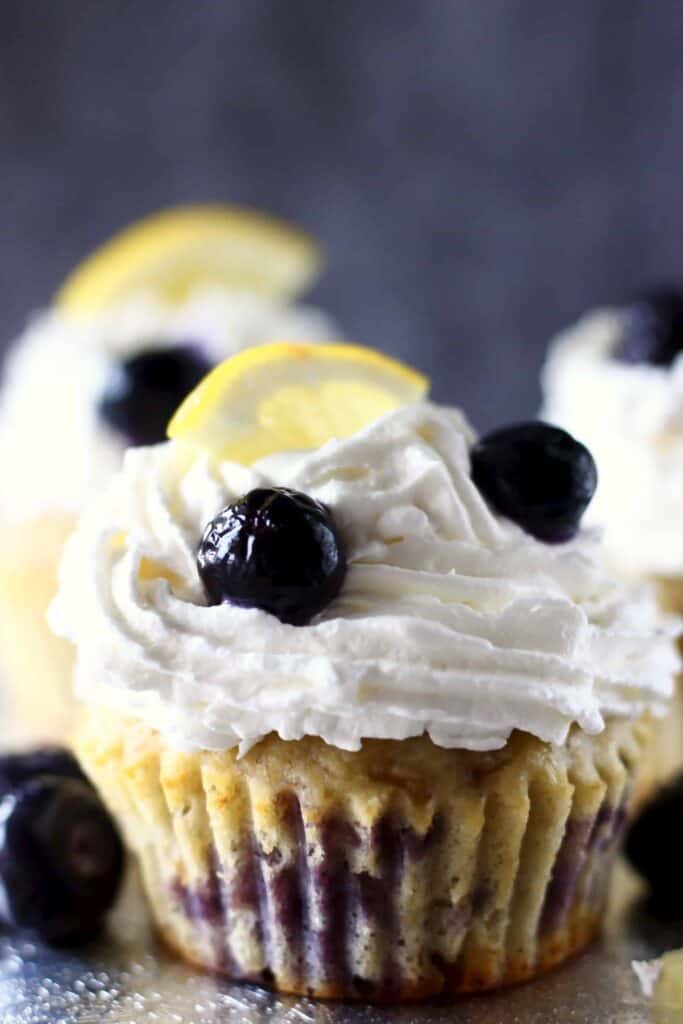 Photo of three blueberry cupcakes topped with white creamy frosting and a fresh blueberry and lemon wedge against a grey background