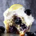 Photo of a blueberry cupcake topped with white creamy frosting and a fresh blueberry and lemon wedge against a grey background