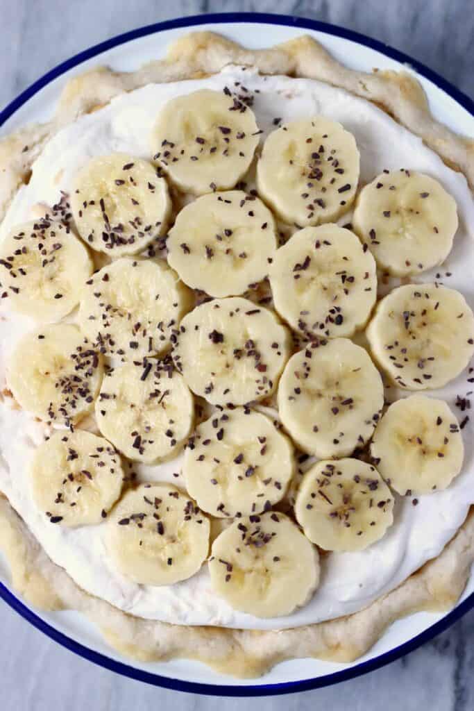 Photo of banana cream pie topped with slices of banana in a blue rimmed pie dish against a marble background