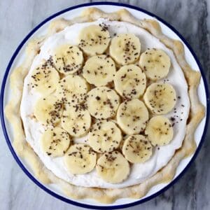 Photo of banana cream pie topped with slices of banana in a blue rimmed pie dish against a marble background