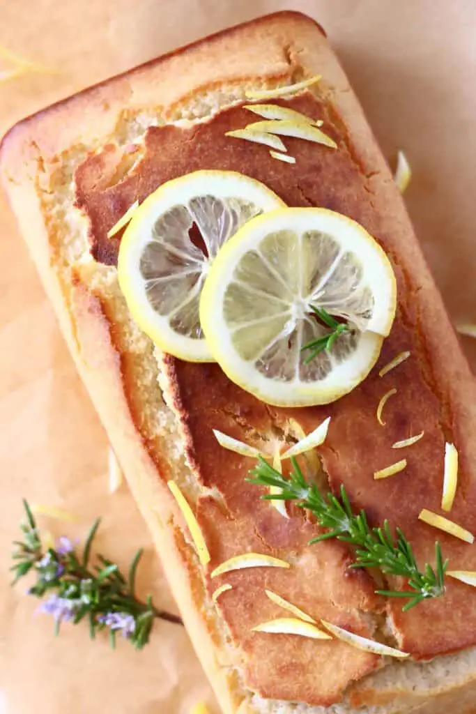 Photo of a golden brown pound cake topped with lemon slices and sprigs of rosemary on top of brown baking paper