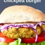 Photo of a curried chickpea burger with a bun, tomato, lettuce and purple cabbage against a grey background