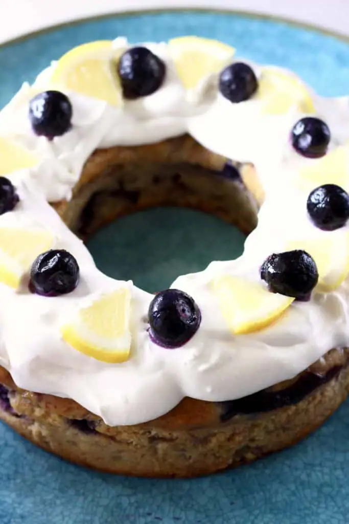 Photo of a bundt cake topped with cream cheese frosting, fresh blueberries and lemon wedges on a big blue plate