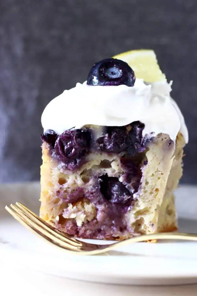 A slice of blueberry bundt cake topped with cream cheese frosting, a fresh blueberry and a lemon wedge on a white plate with a gold fork and a grey background