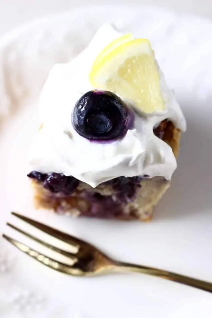 Photo of a slice of bundt cake topped with cream cheese frosting, a fresh blueberry and a lemon wedge on a white plate with a gold fork taken from above