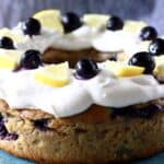 Photo of a bundt cake topped with white frosting, fresh blueberries and lemon wedges against a grey background