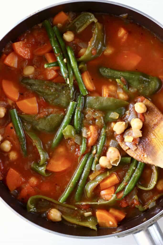 This Vegan Spanish Chickpea Stew is super easy to make, healthy yet satisfying, and packed with veggies! The perfect quick yet nutritious one-pot, plant-based dinner. Gluten-free. #vegan #glutenfree #vegetarian #dairyfree Photo of chickpea and vegetable tomato stew in a saucepan with a wooden spatula holding up some chickpeas