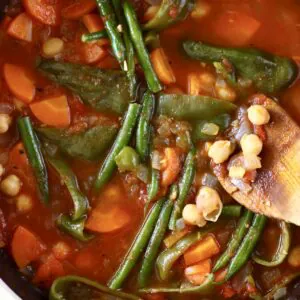 Photo of chickpea and vegetable tomato stew in a saucepan with a wooden spatula holding up some chickpeas