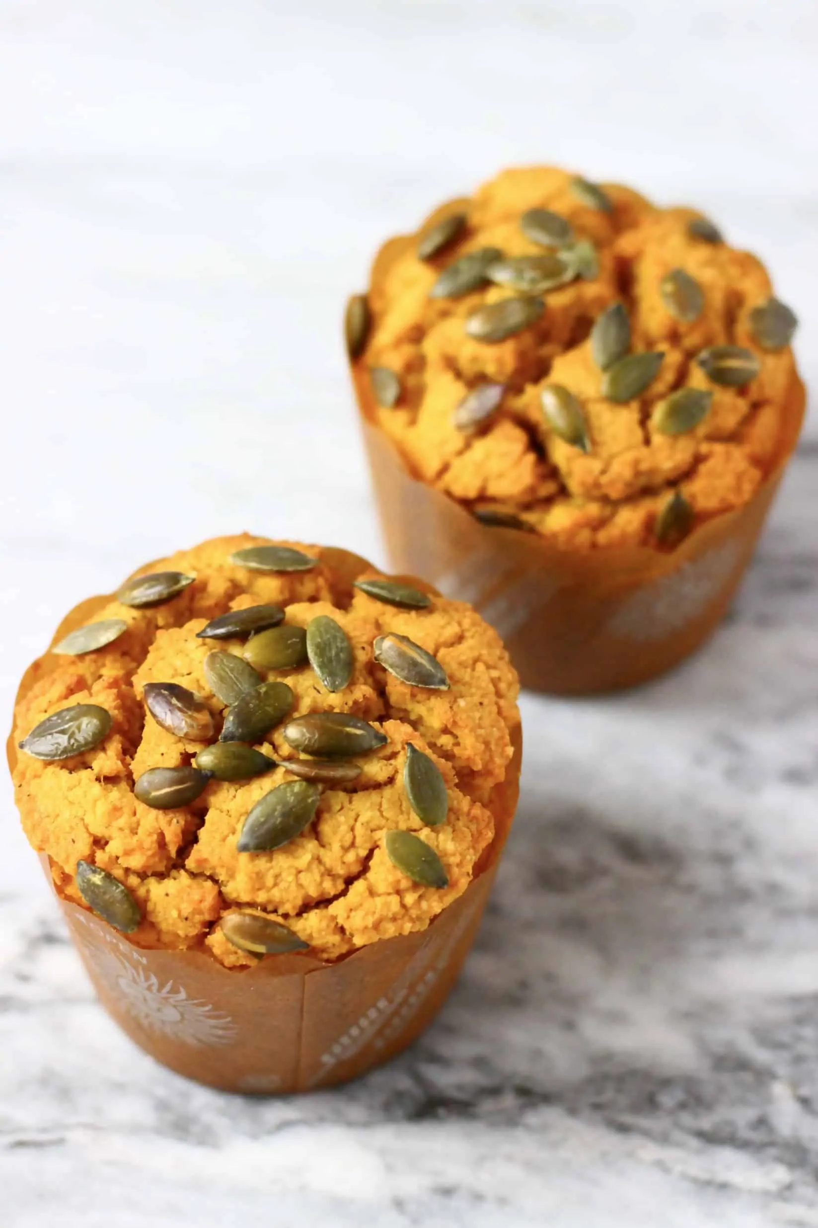 Two gluten-free vegan pumpkin muffins in muffin cases topped with pumpkin seeds