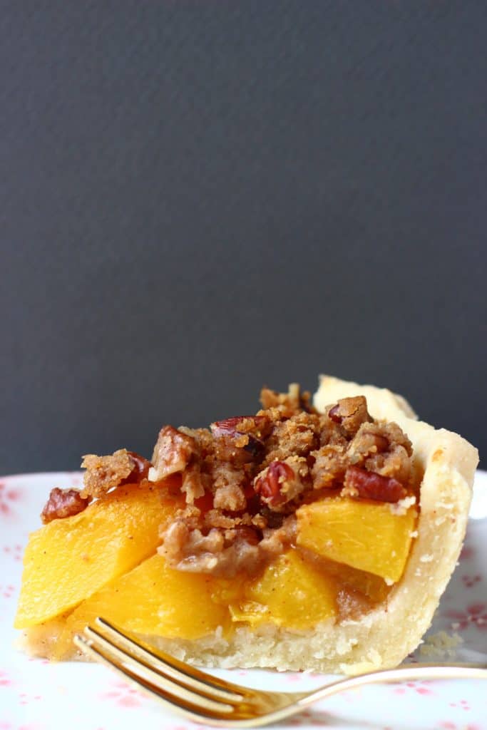 Photo of a slice of peach pie on a white plate with a gold fork and a grey background
