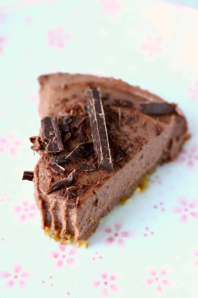 Photo of a slice of chocolate cheesecake topped with dark chocolate shavings on a white plate covered in small pink flowers