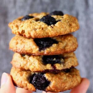 Four blueberry cookies stacked up on top of each other and held up with a hand against a grey background