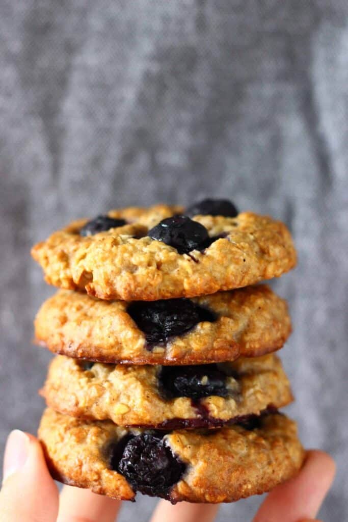 Four blueberry cookies stacked up on top of each other and held up with a hand against a grey background 