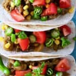 Photo of three tacos on a blue plate filled with roasted eggplant, sweetcorn, sliced green chilli, chopped tomatoes