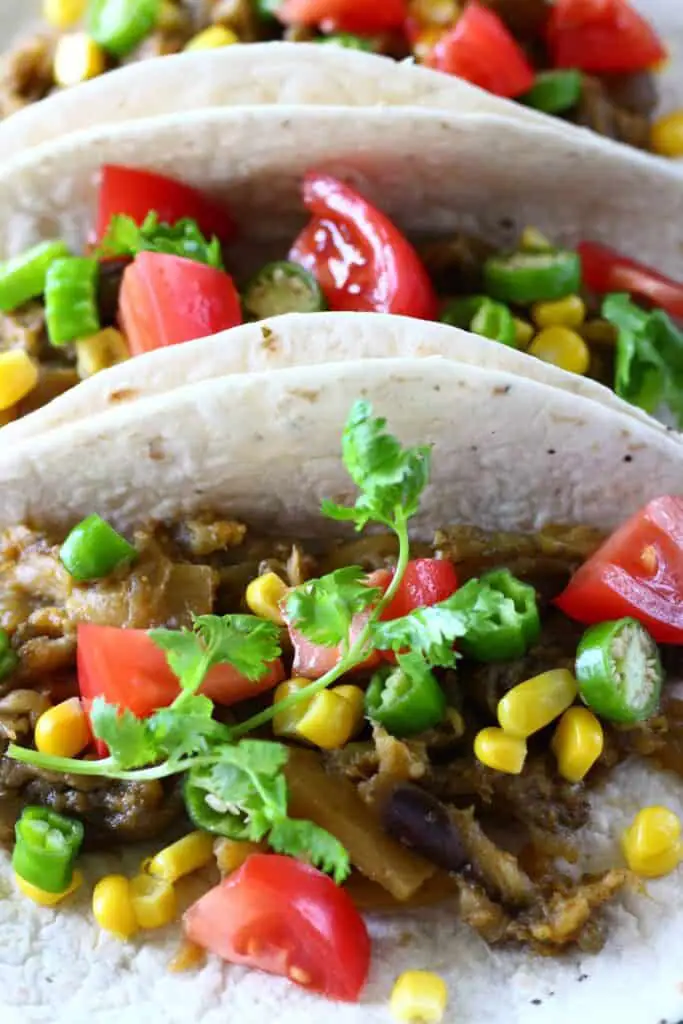 Photo of three tacos on a blue plate filled with roasted eggplant, sweetcorn, sliced green chilli, chopped tomatoes
