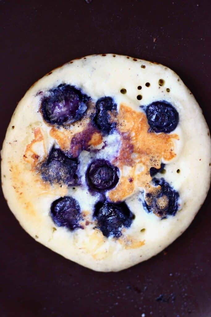 These Gluten-Free Vegan Blueberry Pancakes are super moist, fluffy, perfectly sweet and fruity. They're made in one bowl, and made using simple ingredients without egg replacements, flax eggs or aquafaba. They're egg-free, dairy-free and refined sugar free. They're great for breakfast, brunch, dessert or a snack. #vegan #dairyfree #glutenfree #blueberry #pancakes #veganpancakes #breakfast #brunch #dessert #refinedsugarfree Photo of a blueberry pancake in a frying pan