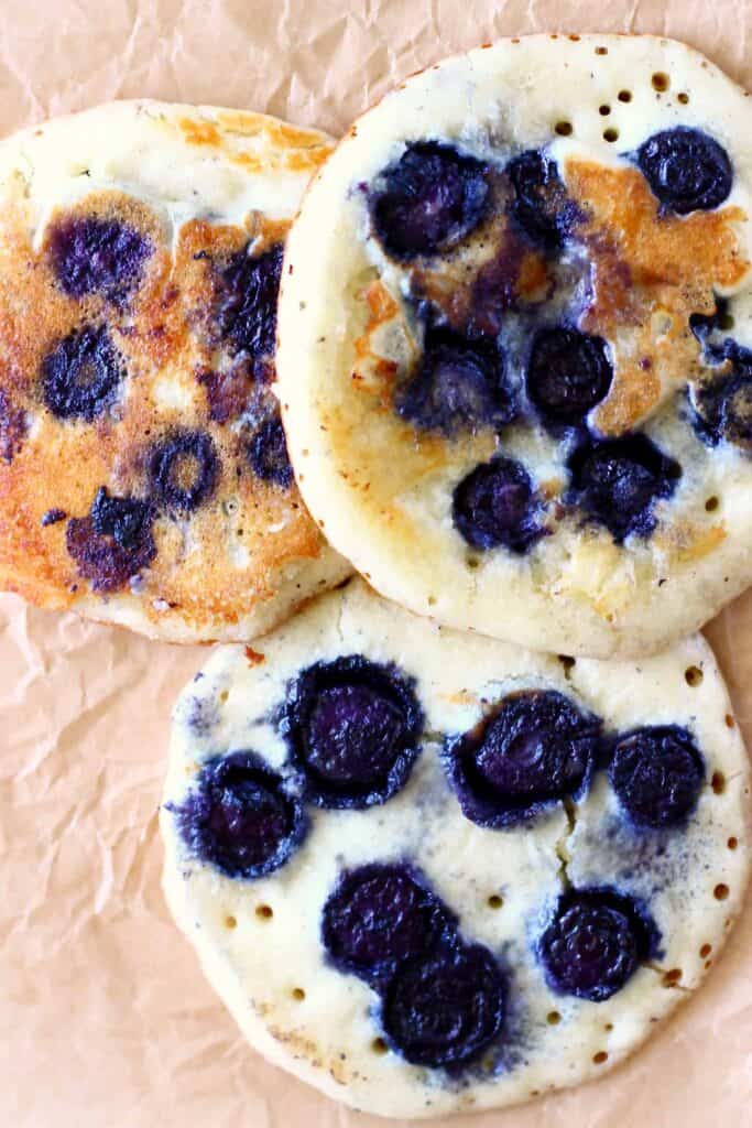 These Gluten-Free Vegan Blueberry Pancakes are super moist, fluffy, perfectly sweet and fruity. They're made in one bowl, and made using simple ingredients without egg replacements, flax eggs or aquafaba. They're egg-free, dairy-free and refined sugar free. They're great for breakfast, brunch, dessert or a snack. #vegan #dairyfree #glutenfree #blueberry #pancakes #veganpancakes #breakfast #brunch #dessert #refinedsugarfree Photo of three blueberry pancakes on a sheet of brown baking paper