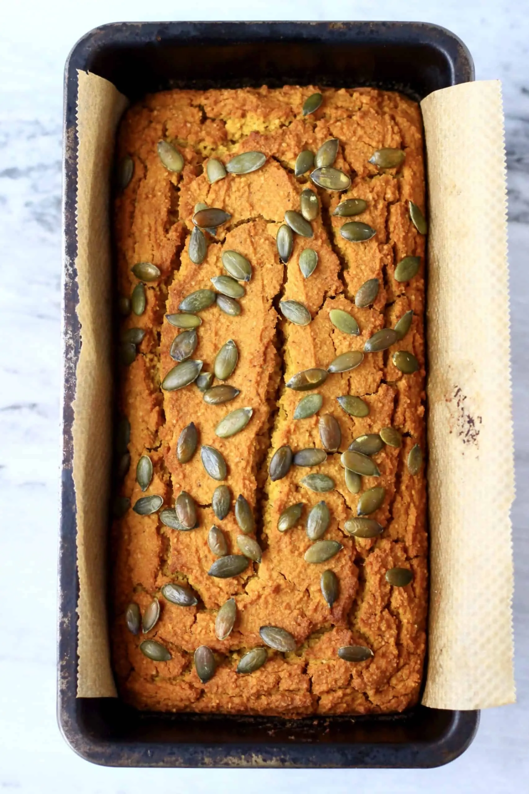 A loaf of gluten-free vegan pumpkin bread topped with pumpkin seeds in a loaf tin lined with baking paper