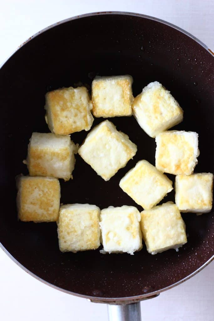 Golden brown cubes of tofu in a dark brown frying pan against a white background