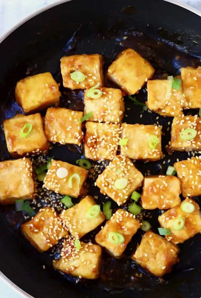 Several pieces of fried tofu covered in sliced spring onions and sesame seeds in a black frying pan