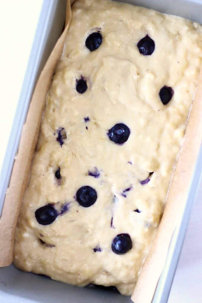 Photo of raw cake batter with blueberries in a silver loaf tin lined with brown baking paper