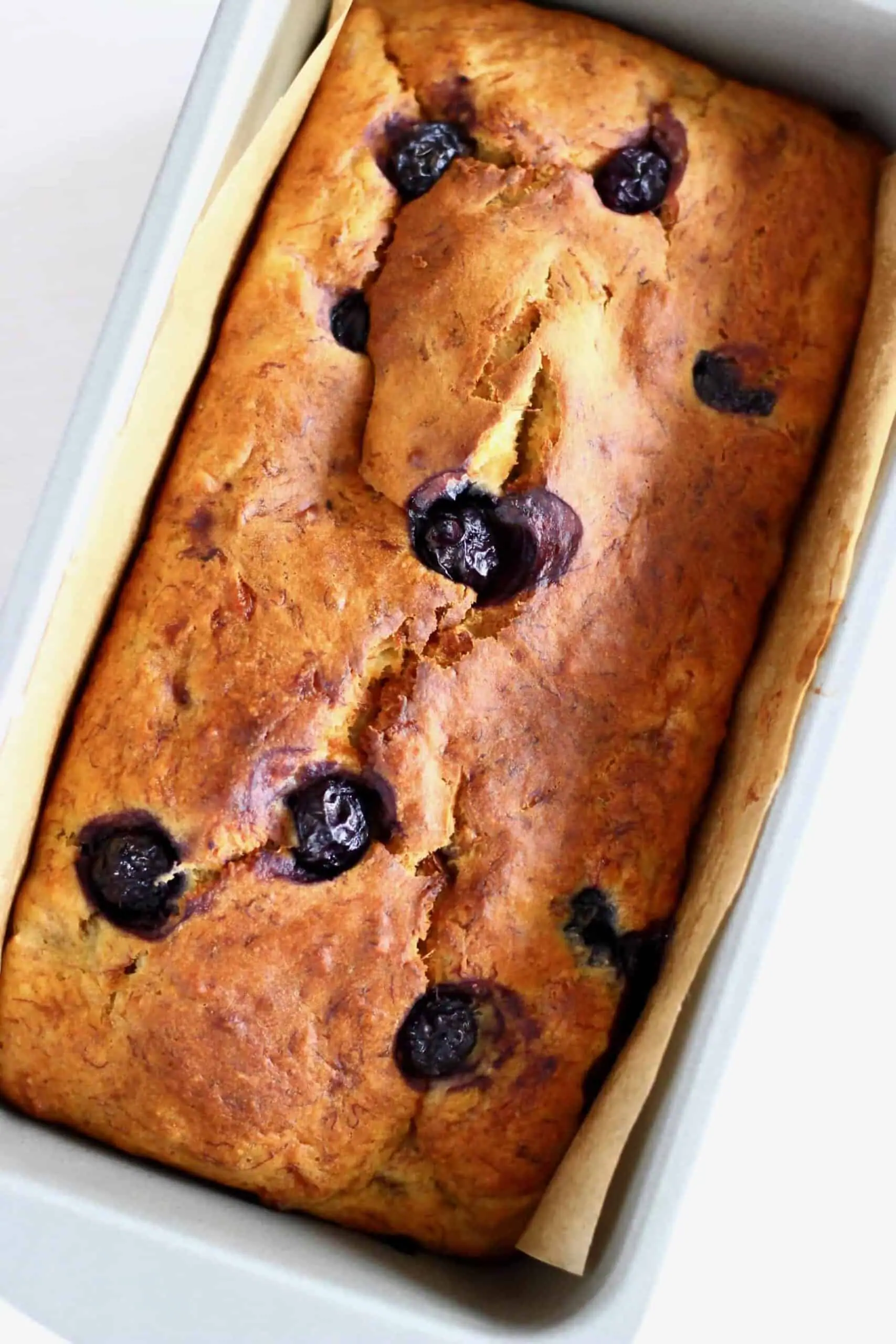 Photo of a golden brown loaf with blueberries in a silver loaf tin lined with brown baking paper