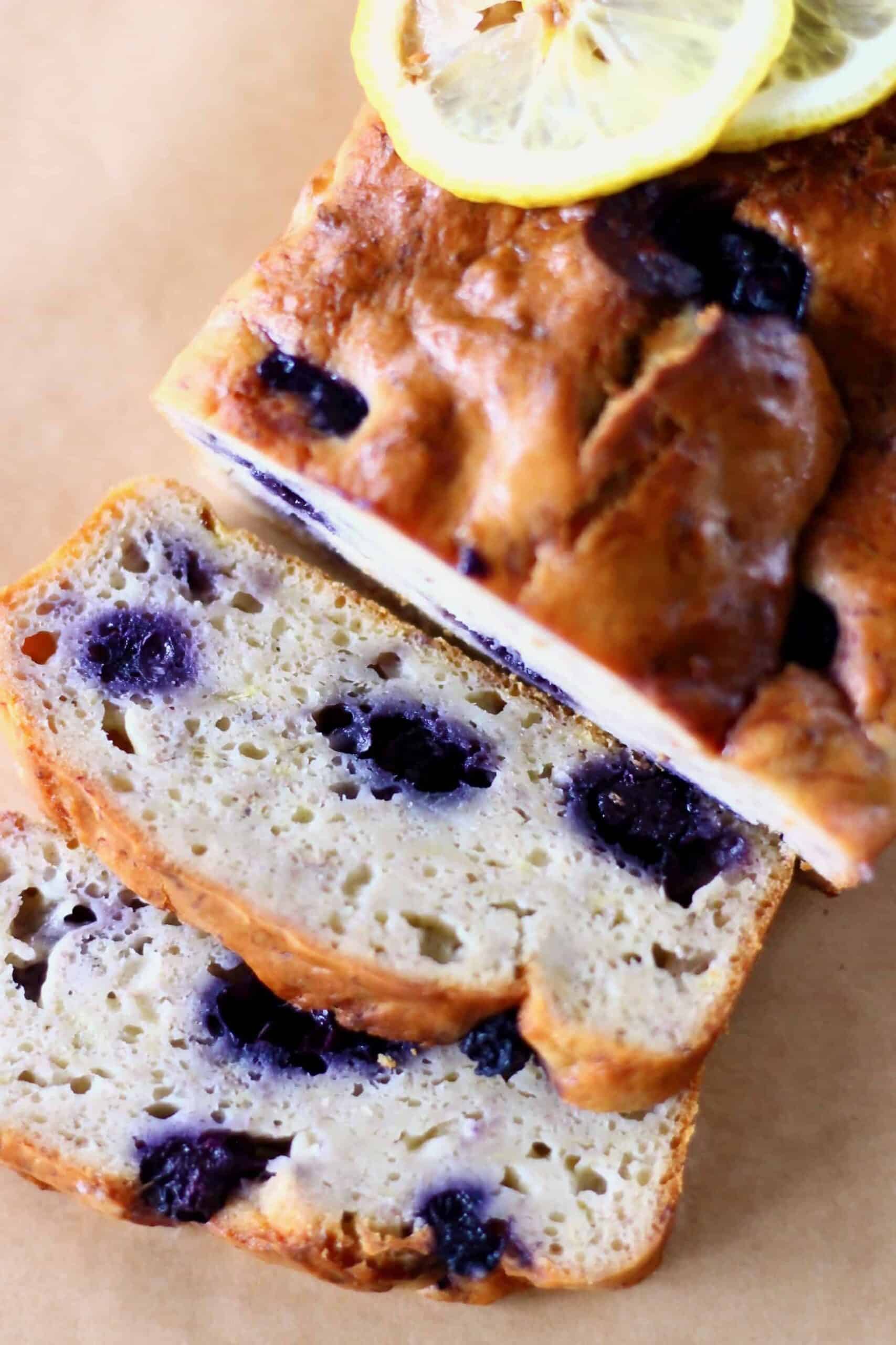This Gluten-Free Vegan Lemon Blueberry Yogurt Cake is seriously moist, bright and citrusy, and packed full of jammy blueberries. It's perfect for dessert, breakfast, brunch or a snack. It's egg-free, dairy-free and refined sugar free. #vegan #glutenfree #dairyfree #blueberry #rhiansrecipes #vegancake Photo of a blueberry loaf cake topped with lemon slices with two slices of it next to it on a piece of brown baking paper