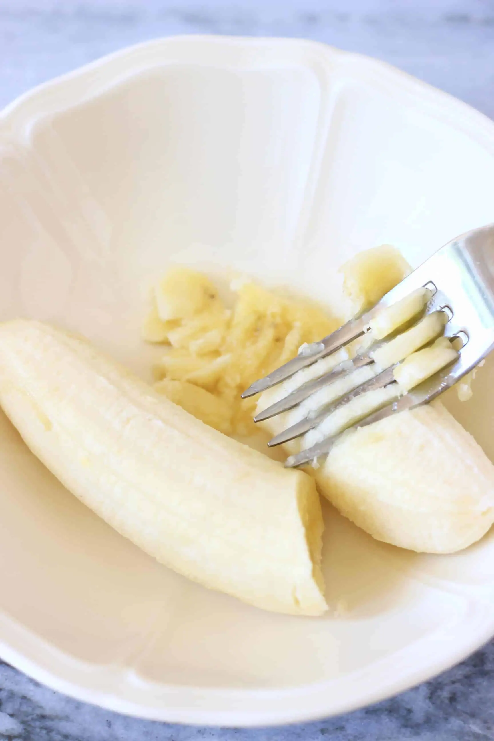 A banana in a white bowl being mashed with a silver fork
