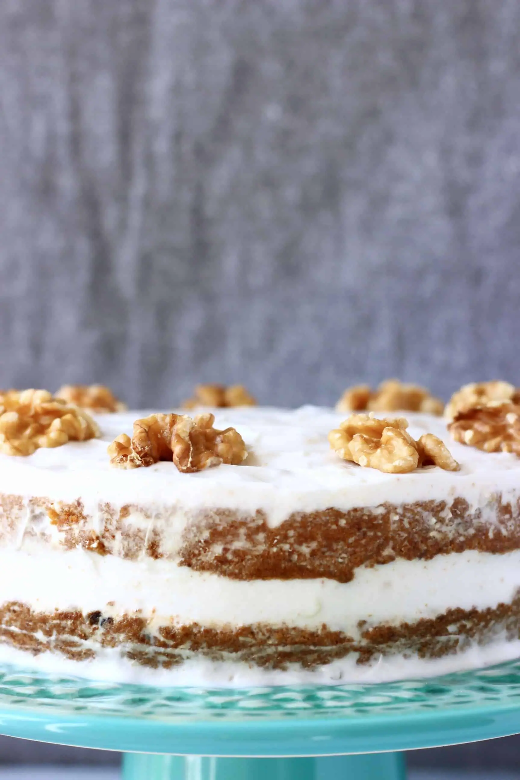 A vegan carrot cake covered in white frosting topped with walnuts against a grey background