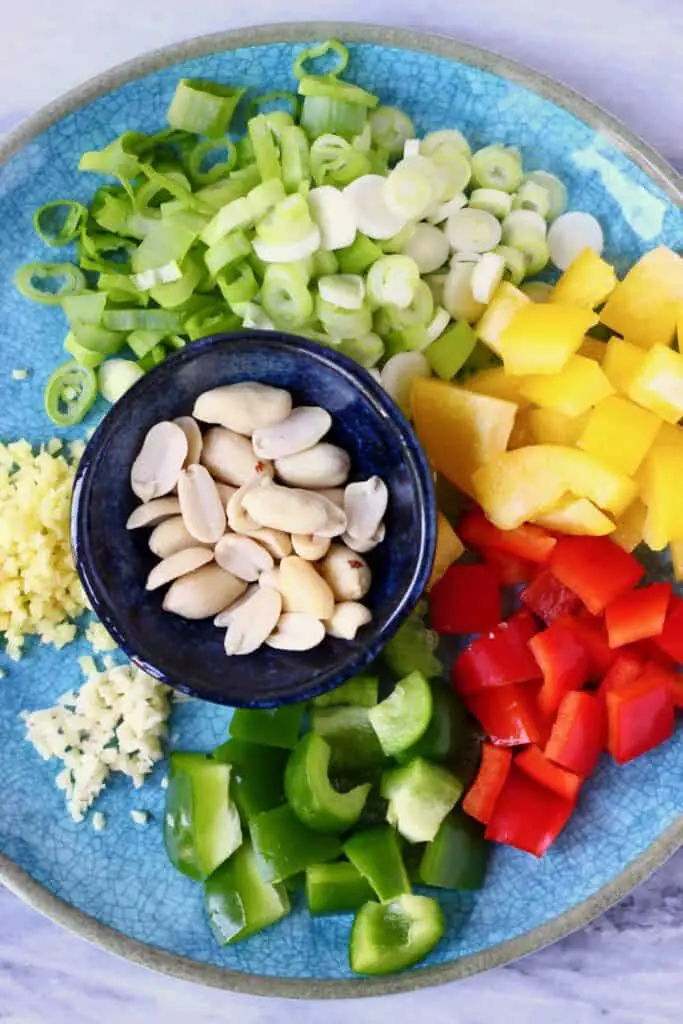 Photo of chopped vegetables and a bowl of roasted peanuts on a blue plate against a marble background