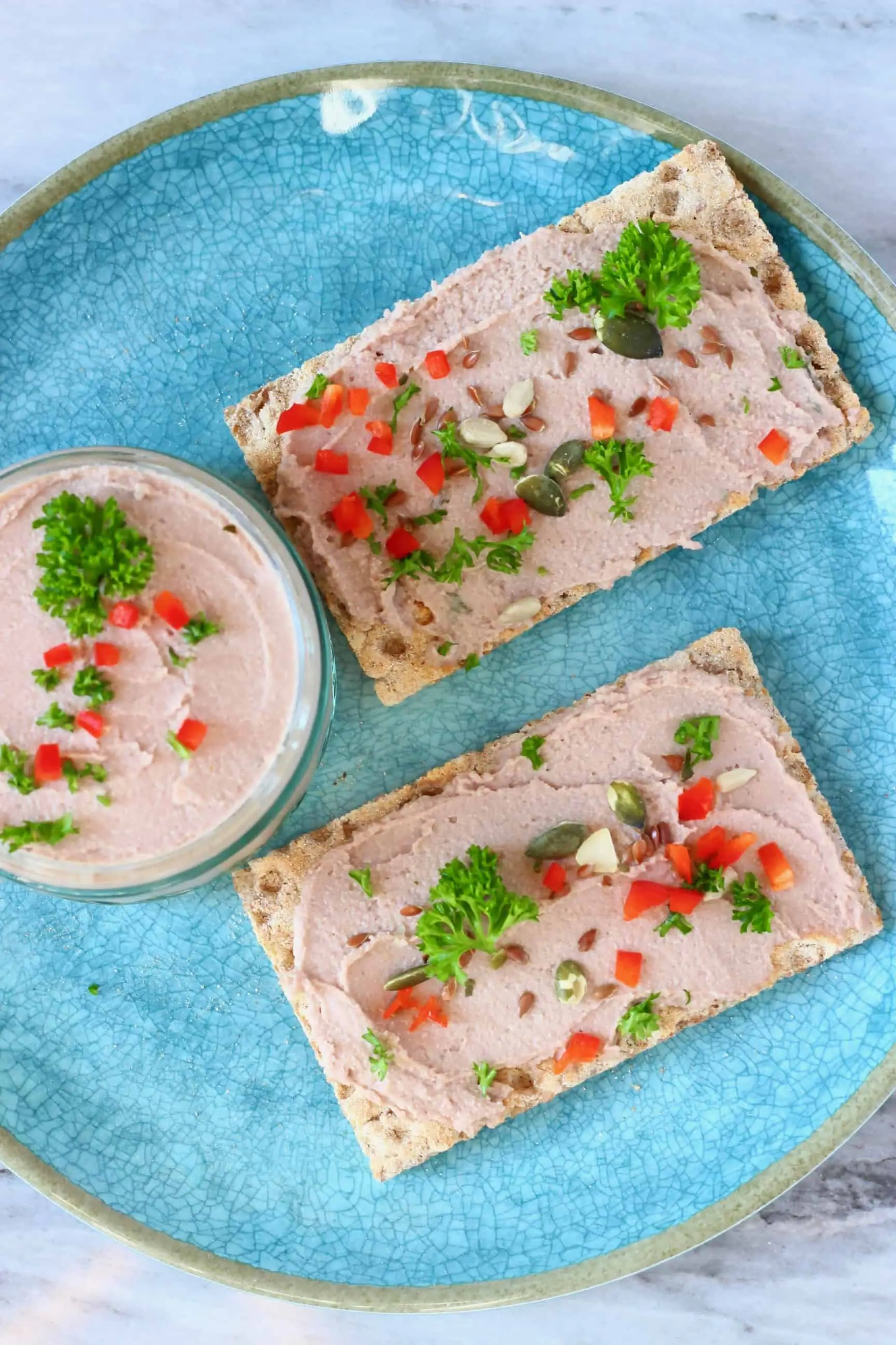 A glass jar filled with vegan mushroom pâté topped with herbs on a blue plate with two rectangular crackers spread with pâté and decorated with pumpkin seeds, chopped red chilli and green herbs