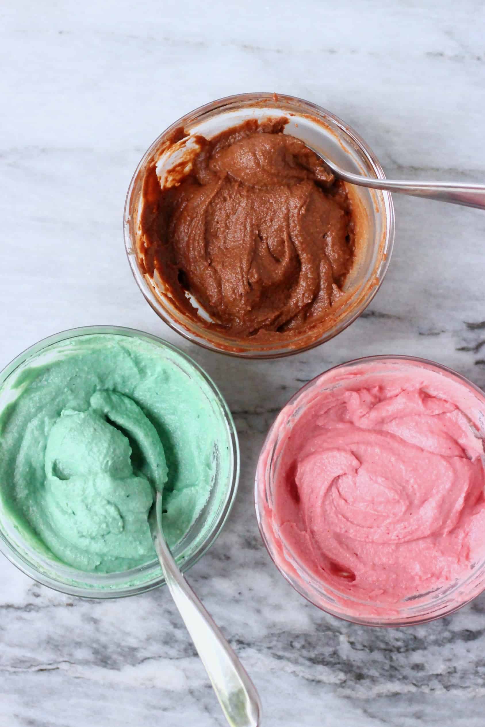 Three bowls with vegan green, brown and pink buttercream frosting on a marble background