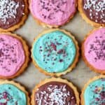 Nine gluten-free vegan sugar cookies topped with different coloured frosting and sprinkles on a sheet of baking paper