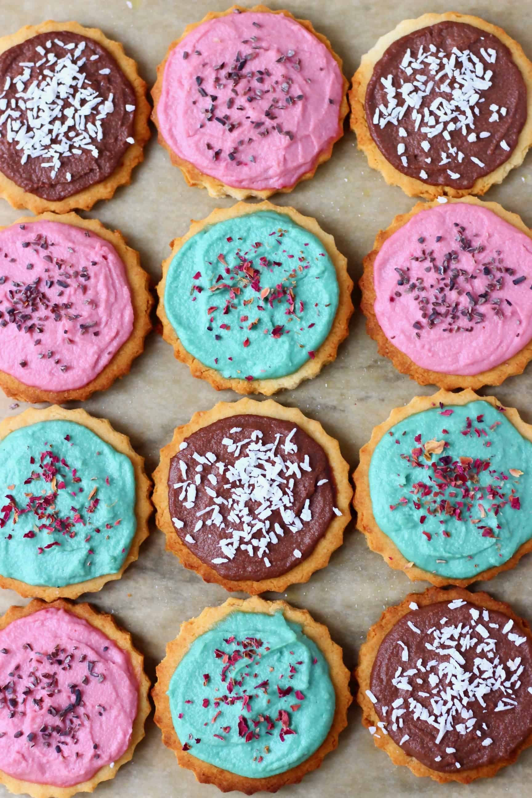Twelve gluten-free vegan sugar cookies topped with different coloured frosting and sprinkles on a sheet of baking paper