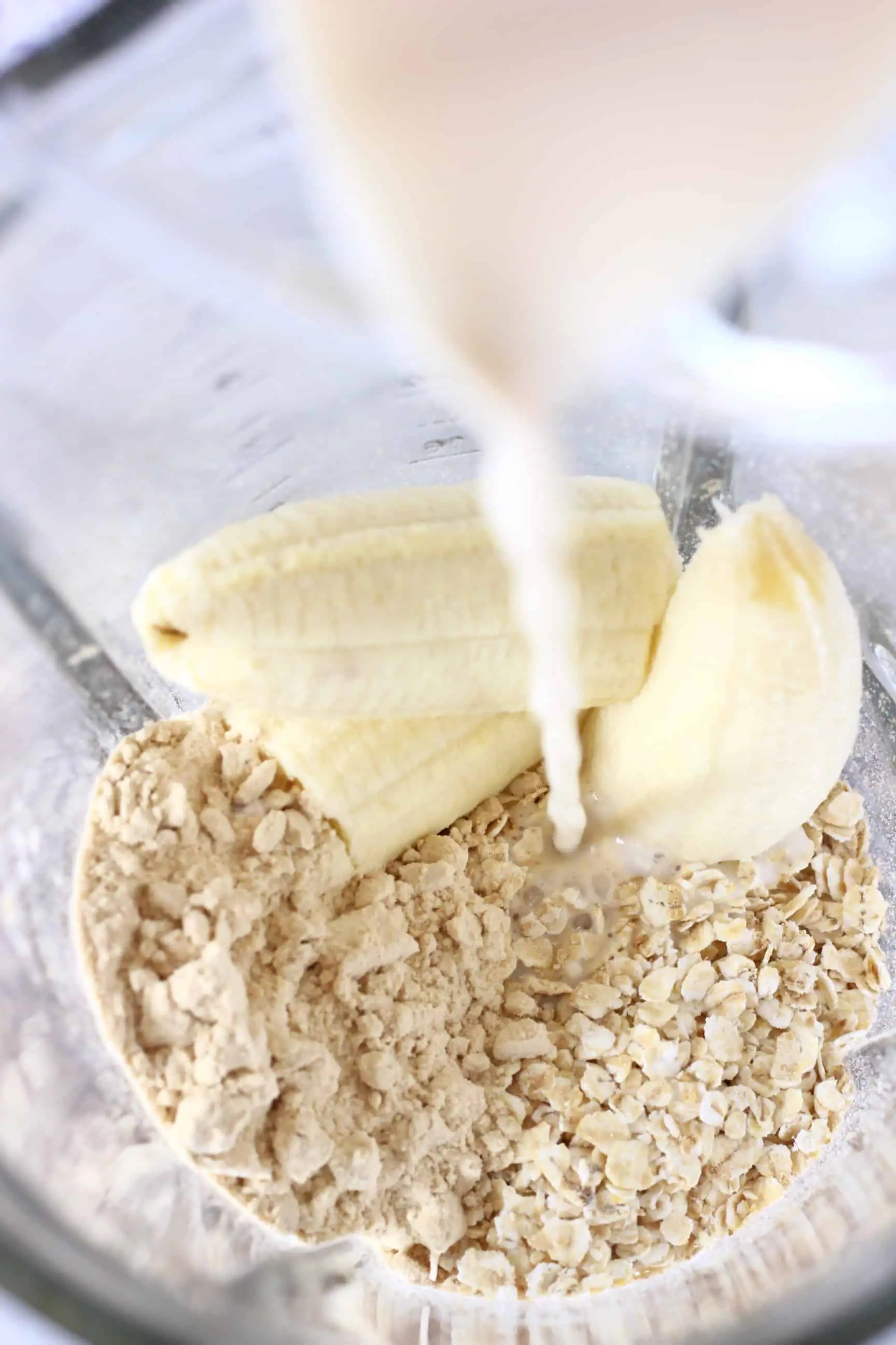 Oats, a banana and protein powder in a glass blender with milk being poured in