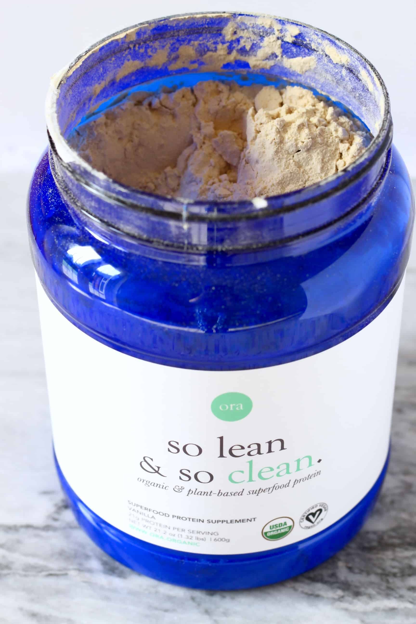 A blue pot of Ora Organic protein powder with the top taken off and brown powder visible inside