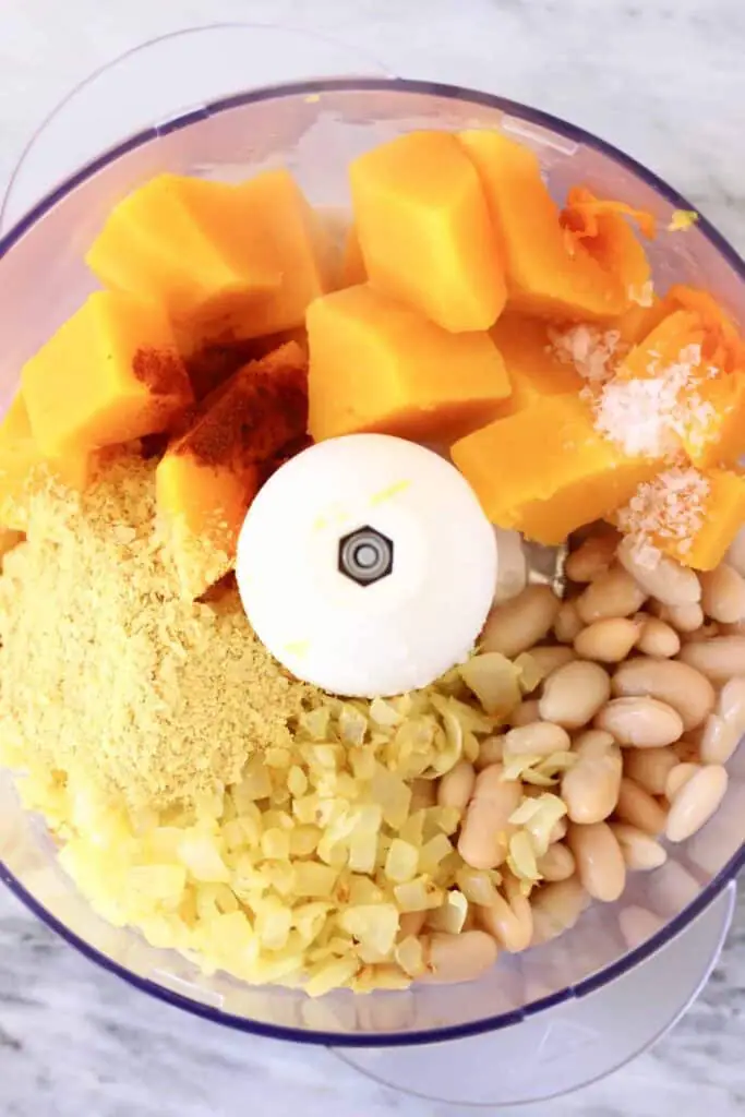 Photo of white beans, cooked butternut squash pieces, nutritional yeast, fried onions and red paprika powder in a food processor against a marble background