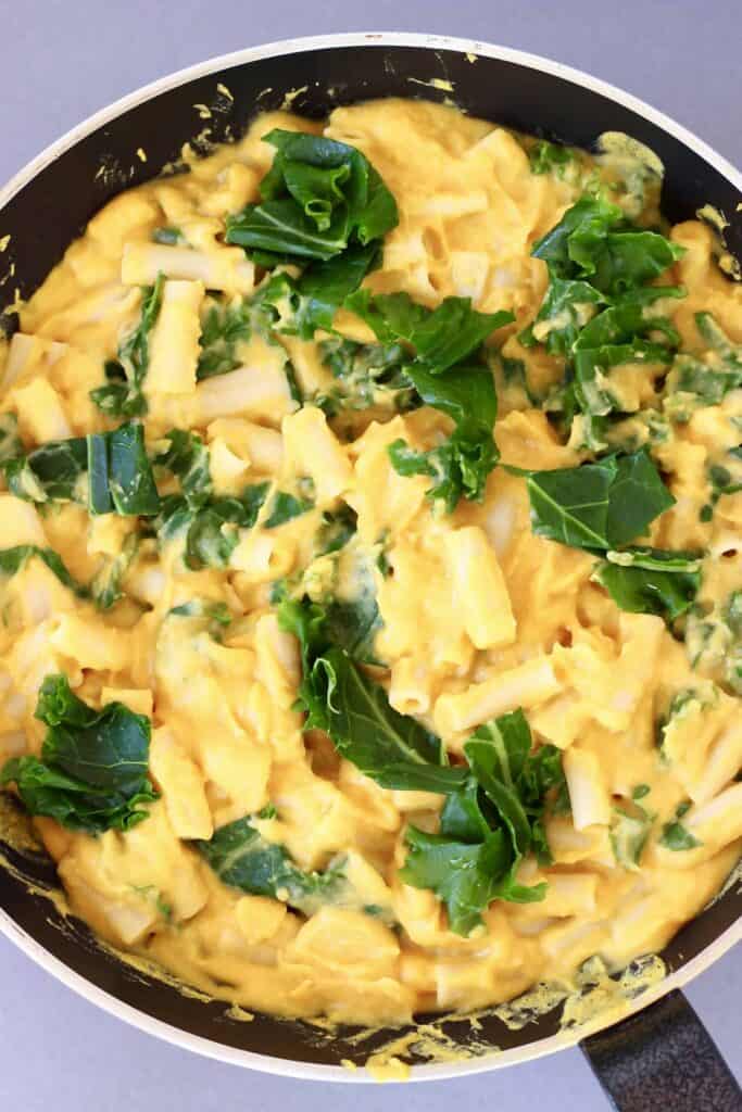 Photo of pasta with orange butternut squash sauce and green kale in a black frying pan against a grey background 