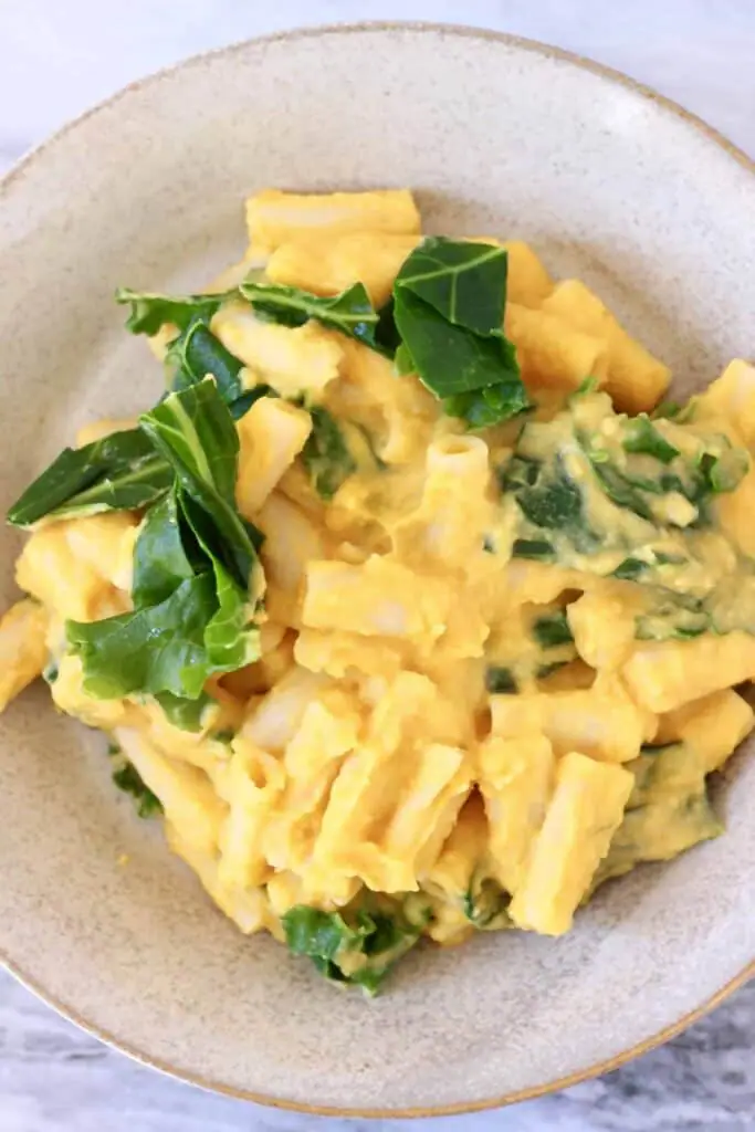 Photo of pasta covered in an orange sauce with green kale in a beige bowl against a marble background