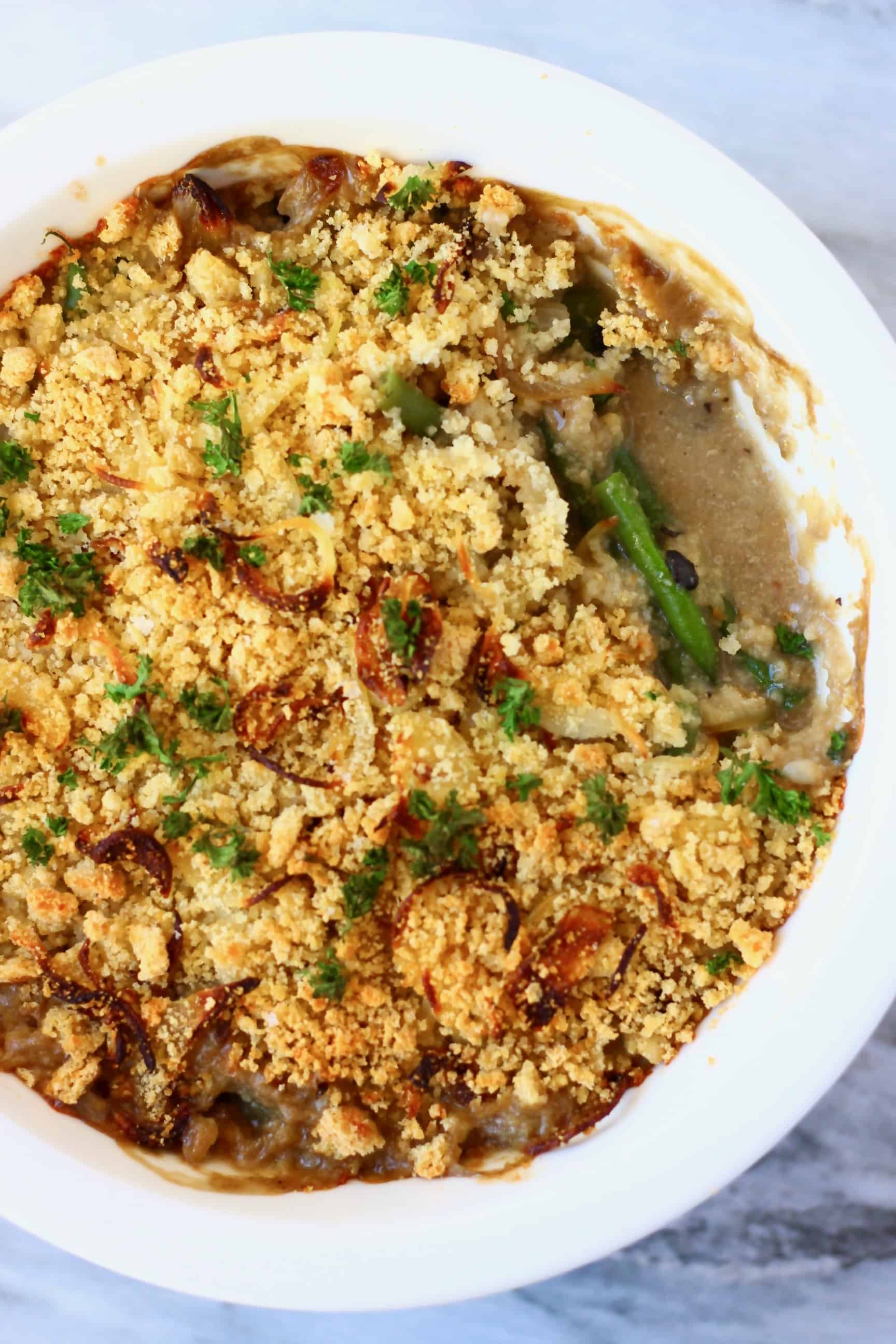 A green bean casserole topped with golden brown breadcrumbs and green parsley in a white pie dish with a mouthful taken out of it 