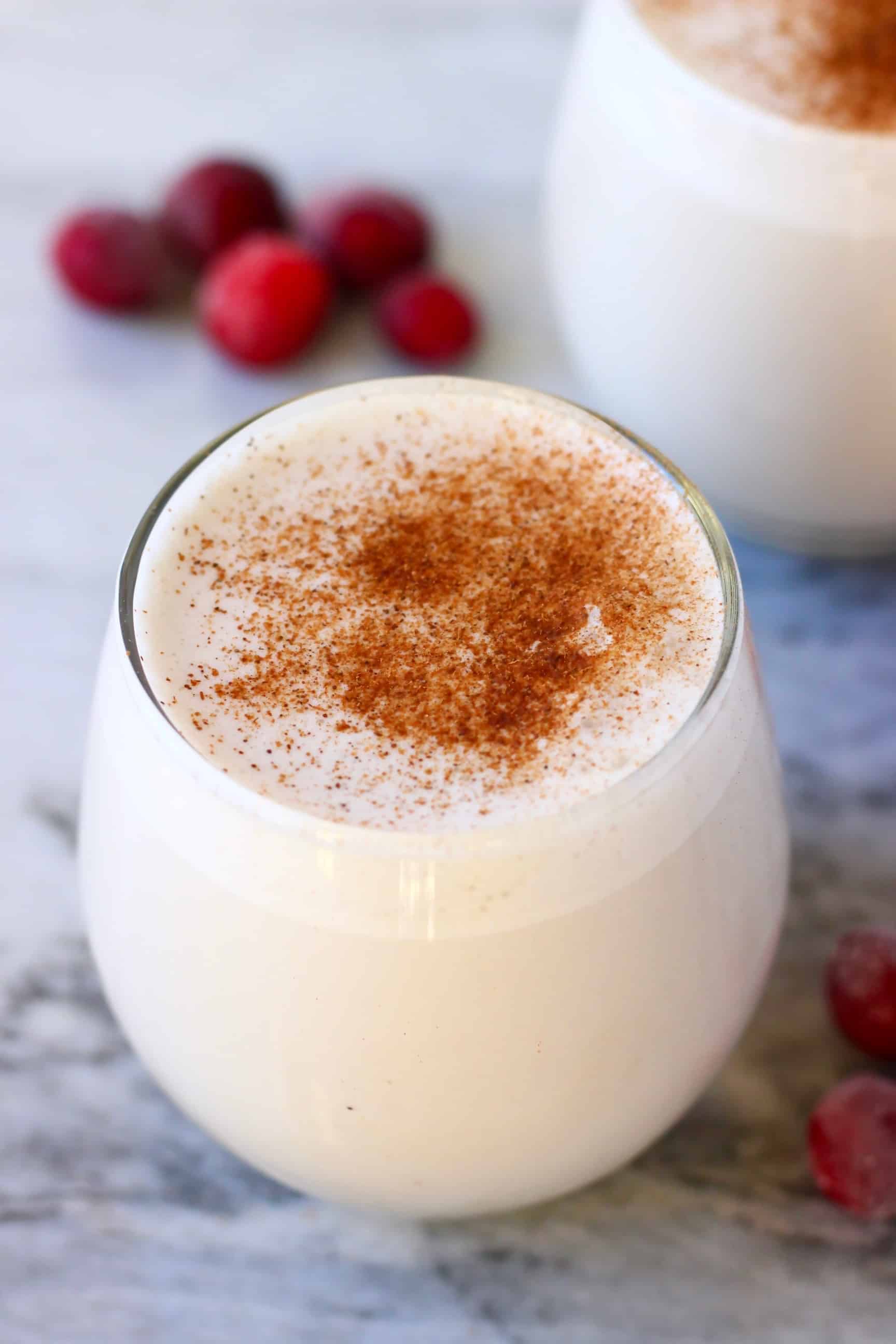 A glass of vegan eggnog topped with cinnamon against a marble background decorated with cranberries