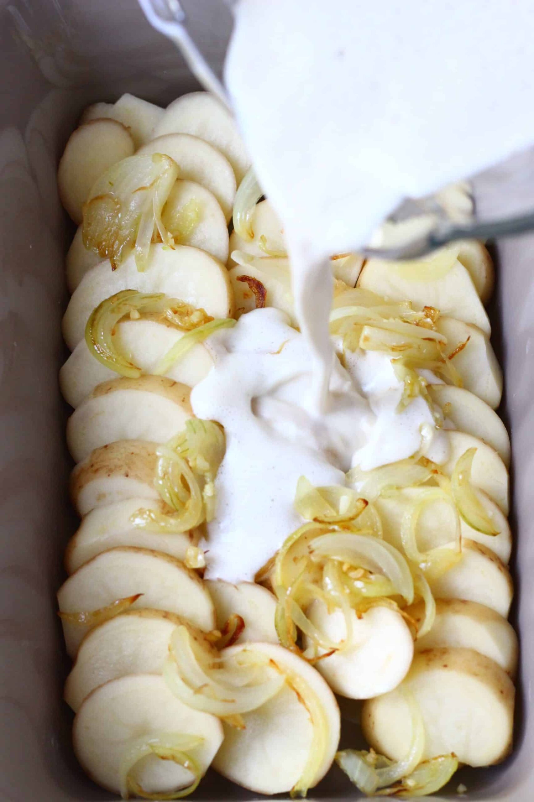 Sliced potatoes topped with fried onions in a baking dish with white cream being poured over the top