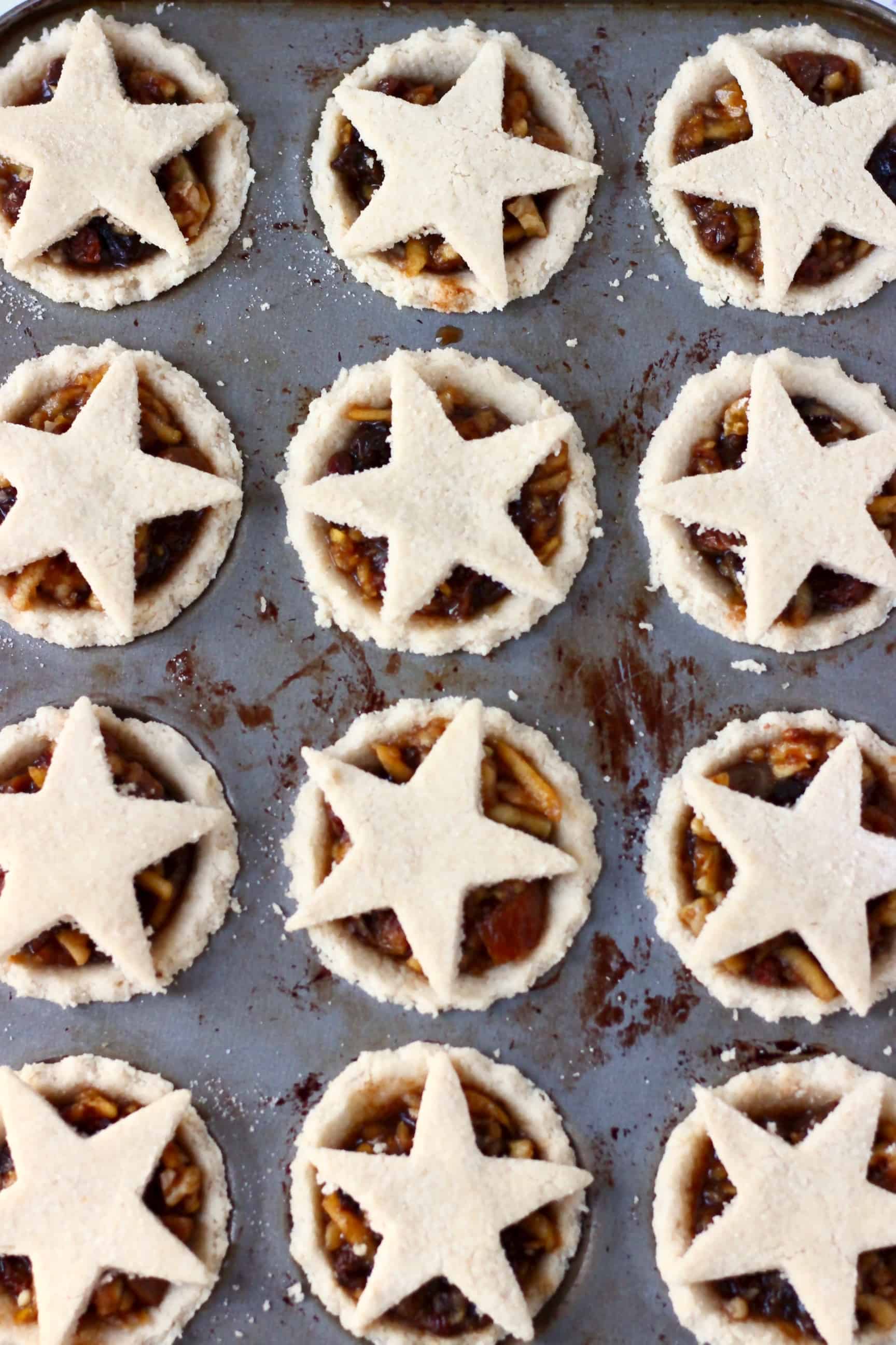 Twelve raw gluten-free vegan mince pies pastry crusts in a muffin tin filled with vegan mincemeat and topped with pastry stars