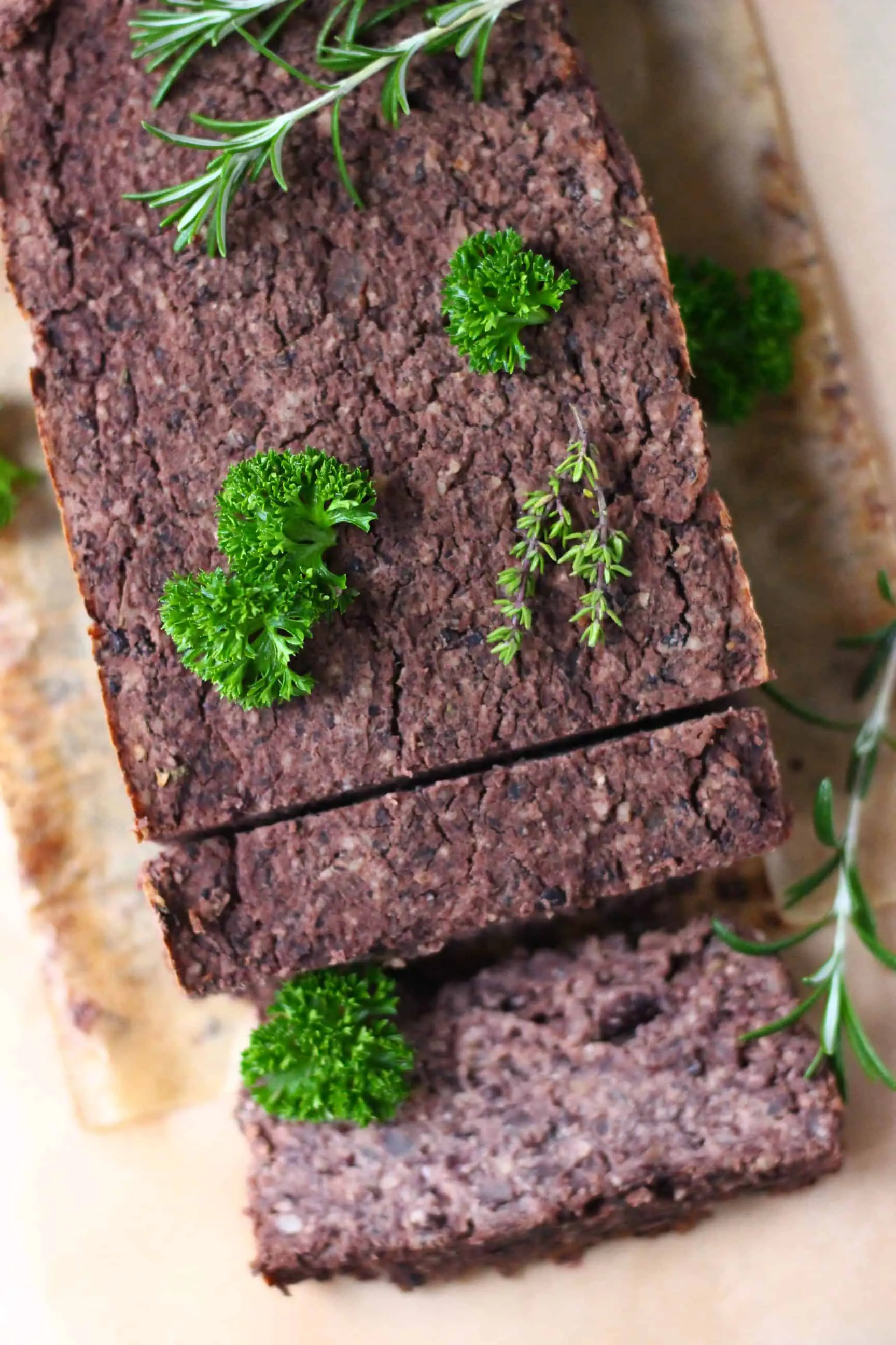 A black meatloaf with two slices taken from it on a sheet of brown baking paper scattered with fresh green herbs