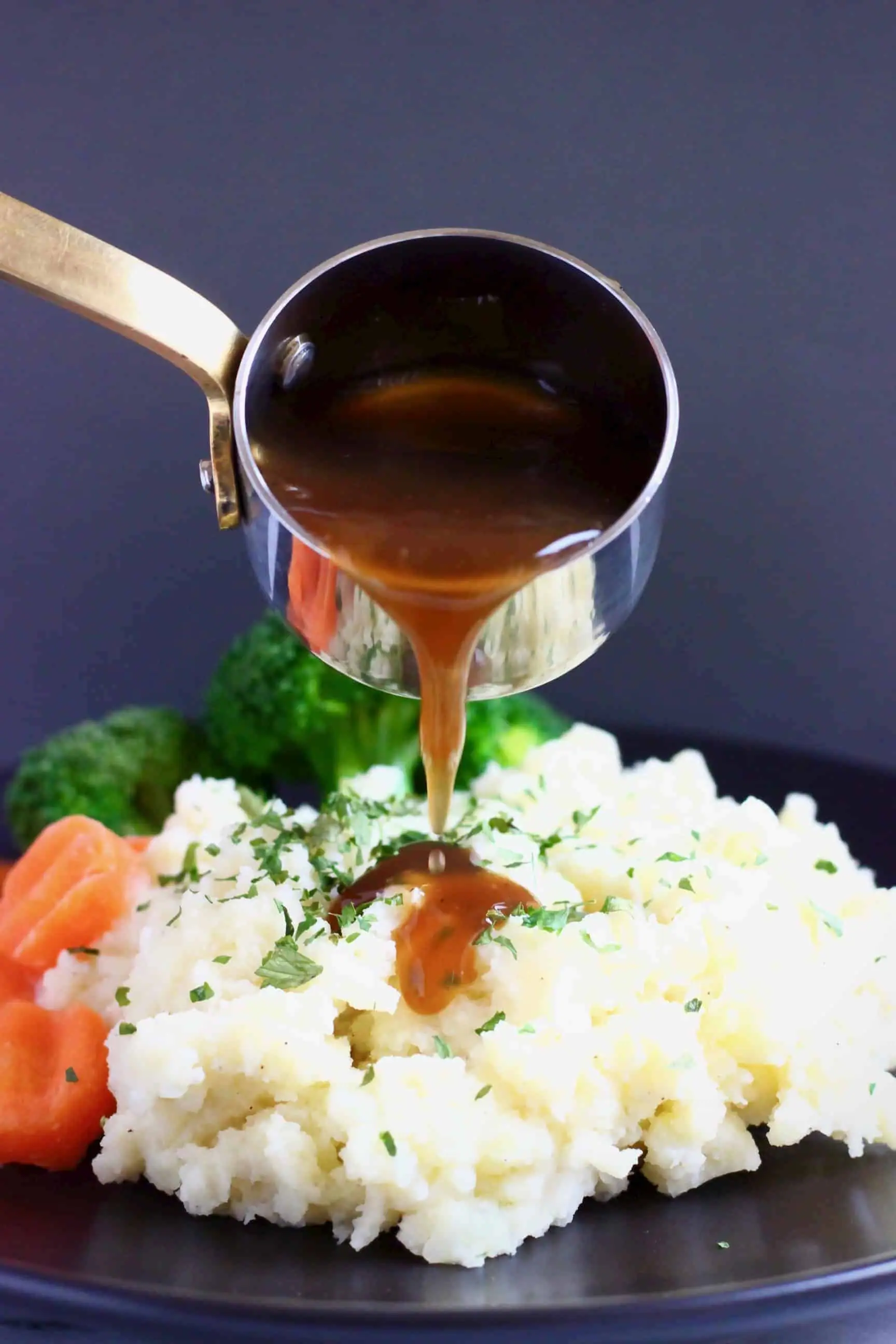A pile of mashed potatoes topped with green herbs with sliced carrots and broccoli on a black plate with brown gravy being poured over in a silver saucepan 