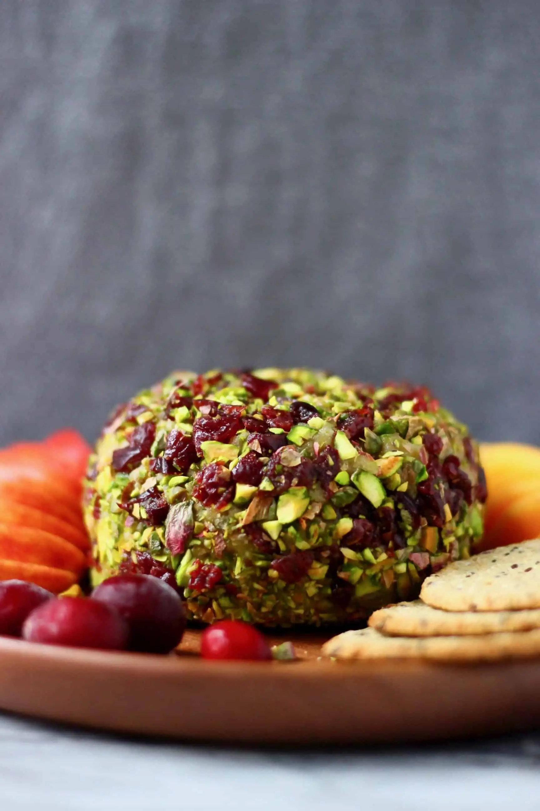 A ball of vegan cashew cheese covered with chopped pistachios and dried cranberries on a wooden plate with sliced apples and oatcakes