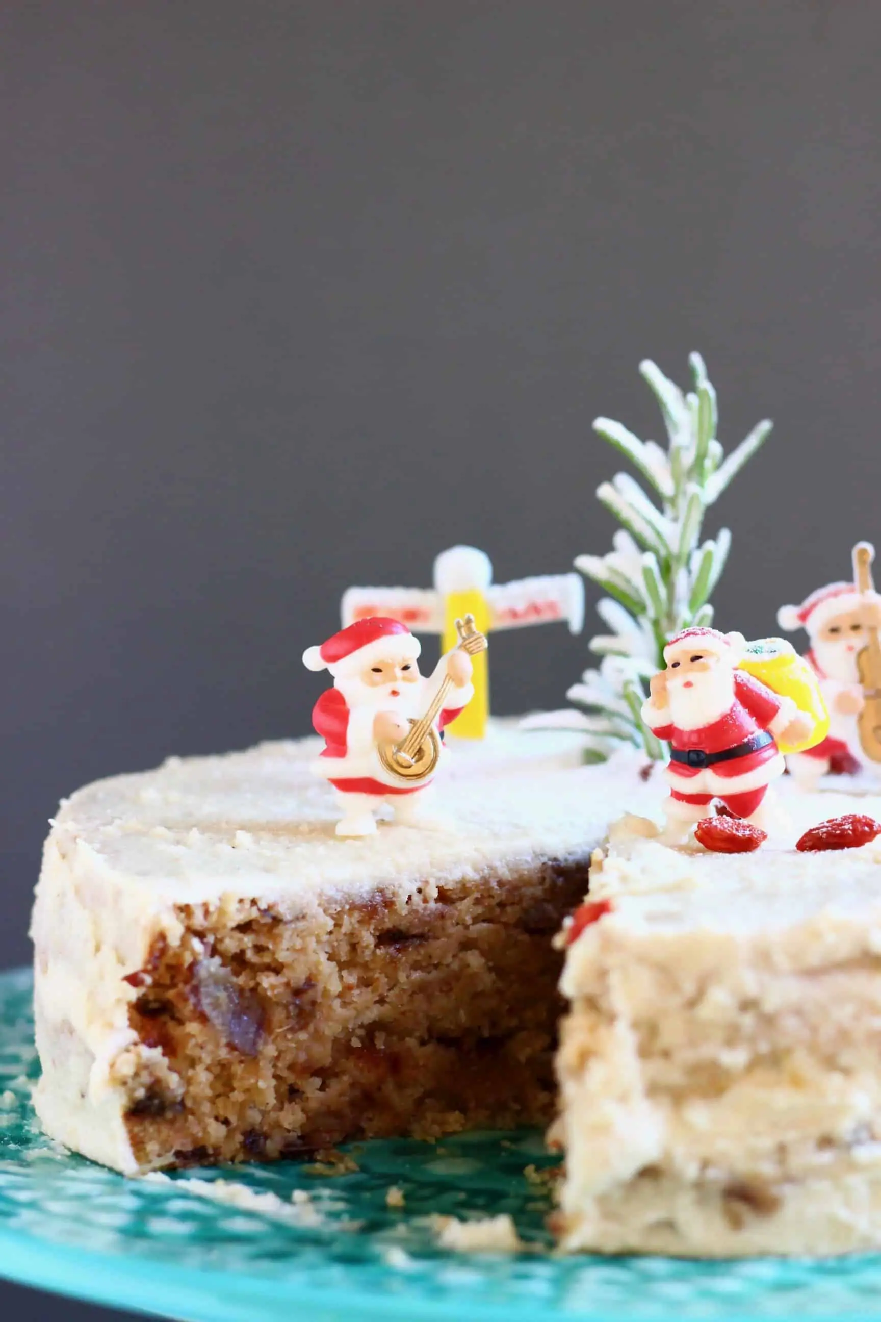 A Christmas fruit cake covered in white buttercream with a slice taken out of it topped with plastic santas against a grey background